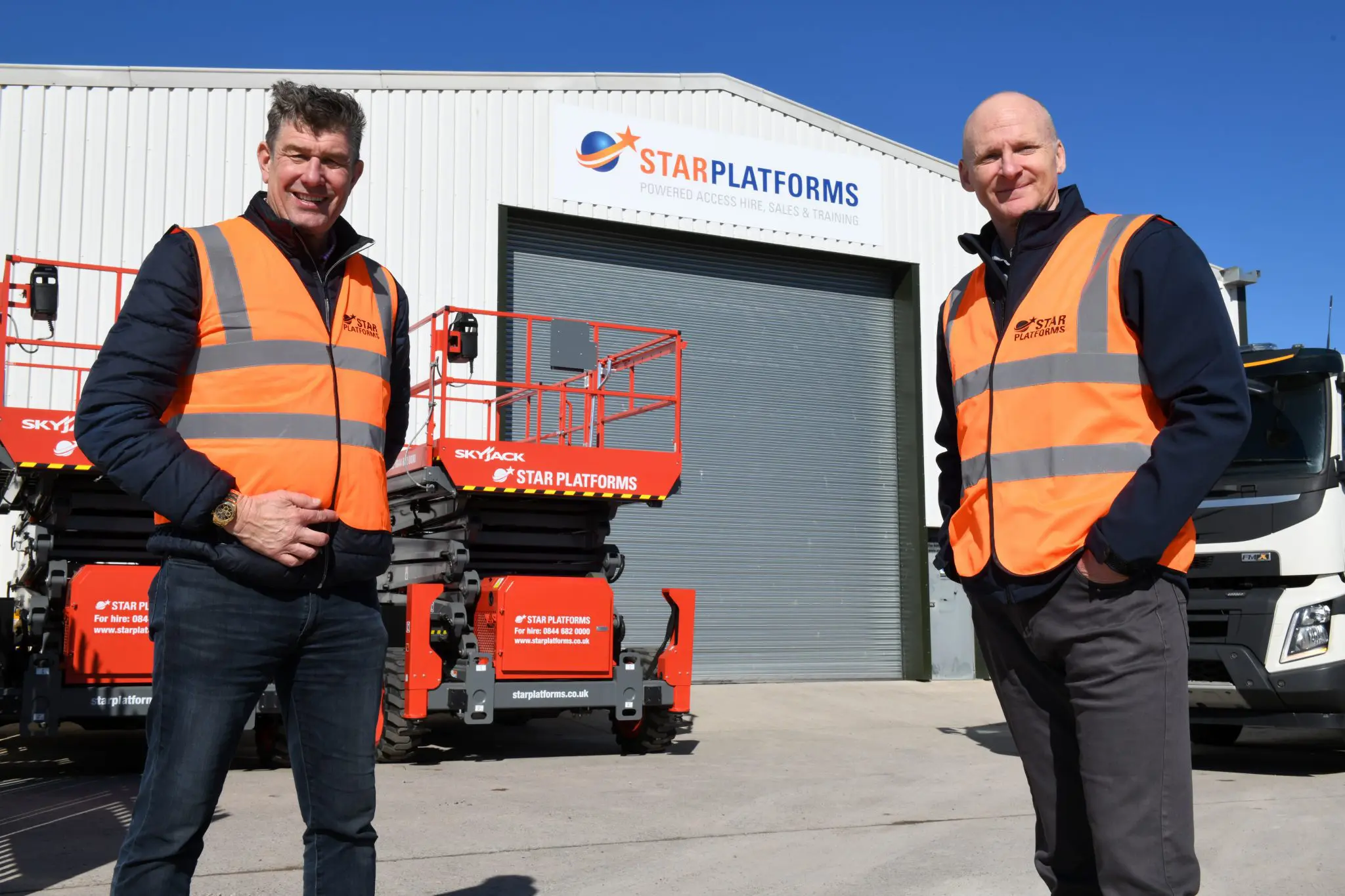 Two white men in high vis vests stand on either side of the image. Behind them is the South West depot building - a white building with a single garage door. Between the men and the building is a scissor lift, parked to the left of the closed garage door.